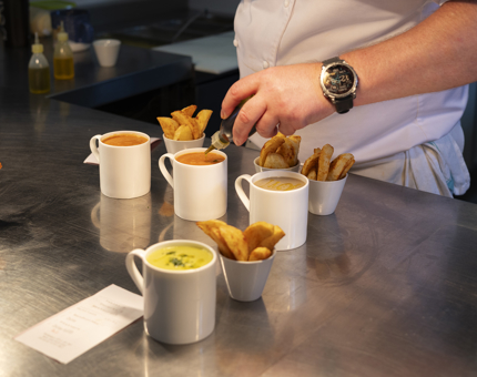 A chef is in the Galley aboard Britannia, putting the finishing touches on mugs of soup and potato wedges. 
