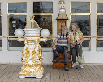 On The Royal Yacht Britannia in Edinburgh a man and woman sit under a Bell. The gold and white decorative binnacle is in the foreground