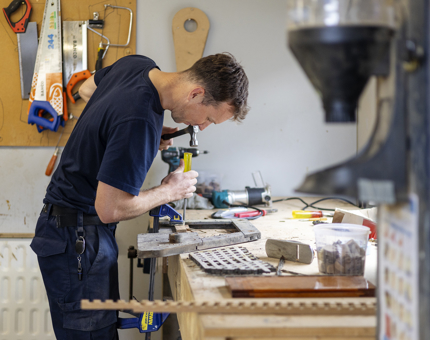 A Maintenance man is standing at a workbench chiseling wood. 