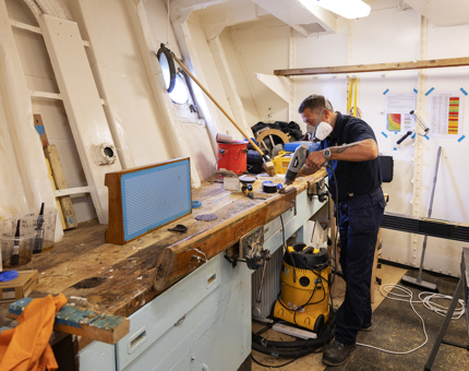 A maintenance man is in the workshop holding a heat gun to remove varnish from a handrail. 