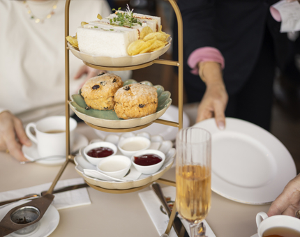 A 3 tiered cake stand with sandwiches, scones and jam. there are cups of tea and fizz on the table, there are two people in the background. 