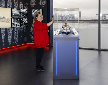 A Housekeeper is polishing a glass cabinet containing a model ship of The Royal Yacht Britannia, through the windows behind her you can see the Port of Leith. 