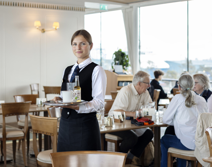 A Hospitality server is standing in the middle of the Royal Deck Tearoom holding a tray of drinks. There are people sitting at tables in the background. 