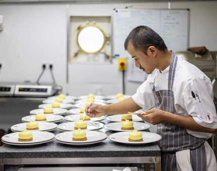 A Chef is in the Galley aboard Britannia adding edible gold leaf to small cheesecakes.