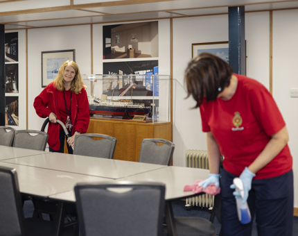 Two Housekeepers are cleaning the onboard Training Room. One is hoovering, the other is wiping the table. There is a model of Britannia in the background. 