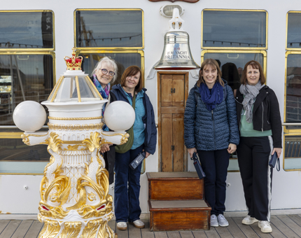 Four women pose for a photo by The Royal Yacht Britannia's Bell and decorative binnacle.