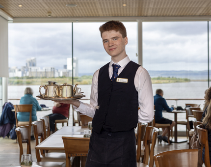 A Hospitality Server is standing in the Royal Deck Tearoom and holding a silver tray with two silver teapots on it. There are guests sitting at tables behind him. 