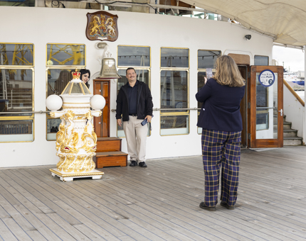 A Visitor Assistant is taking a photo of two visitors by Britannia's Bell on the Verandah Deck. 