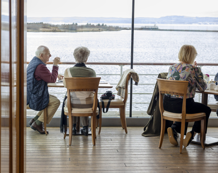 Visitors are sitting at tables in the Royal Deck Tearoom on Britannia enjoying the views of the Firth of Forth. 