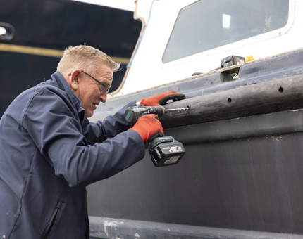 A close up look at a Maintenance team member holding a power drill working to refurbish the Admiral's Fast Motor Launch boat. 
