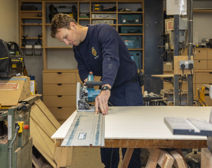 A Maintenance team member is at a work bench cutting a sheet of wood. 
