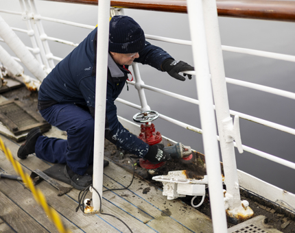 A Maintenance man is kneeling next to the railings on the Verandah Deck, he is using a grinder to remove rust. 