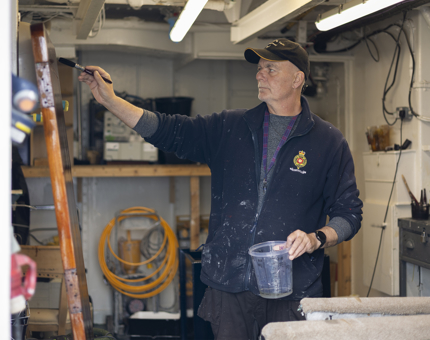 A Maintenance team member holding a varnish pot and paintbrush, he is varnishing a wooden door. 